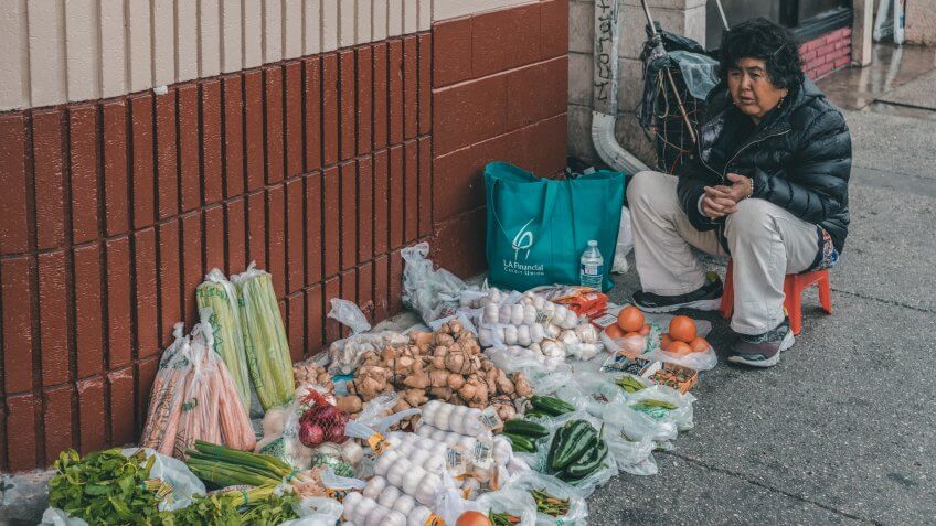 Los Angeles, CA, USA - April 5, 2018: Female street vendor sits along Broadway in Chinatown and sells small amounts of several vegetables and fruits displayed on the surface of the sidewalk.