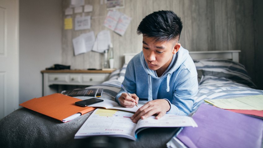 Teenage boy lying on his bed while concentrating on homework for his exams.