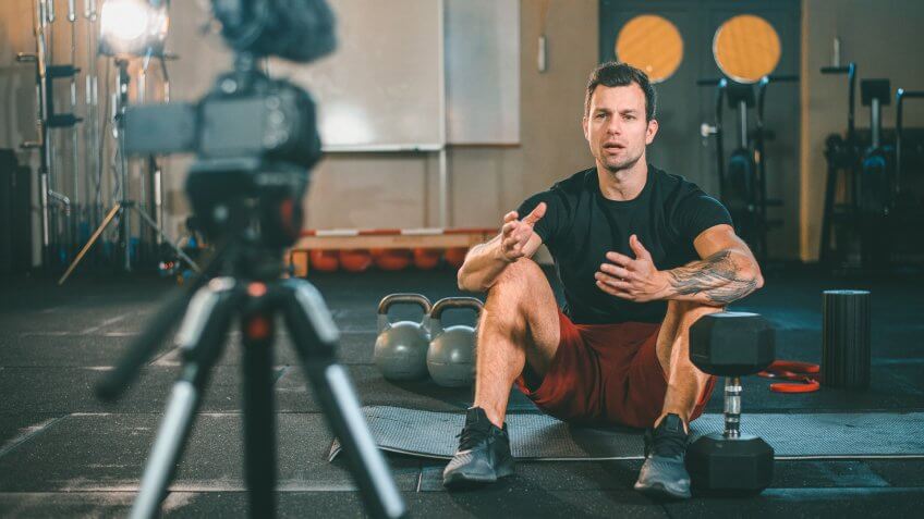 Caucasian male fitness instructor sitting on exercise mat at gym and making vlog explaining how to workout with kettlebells and dumbbells.