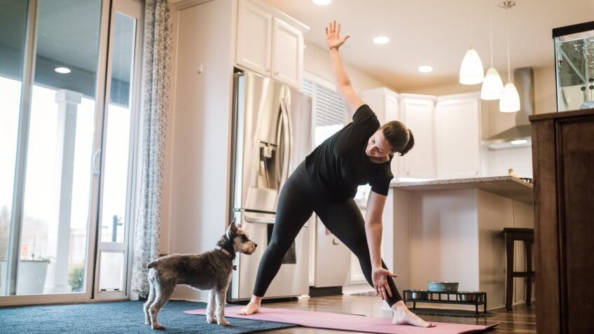 A mature adult woman does yoga and strength training exercises on a mat in her living room, her pet terrier dog keeping her company and trying to play.