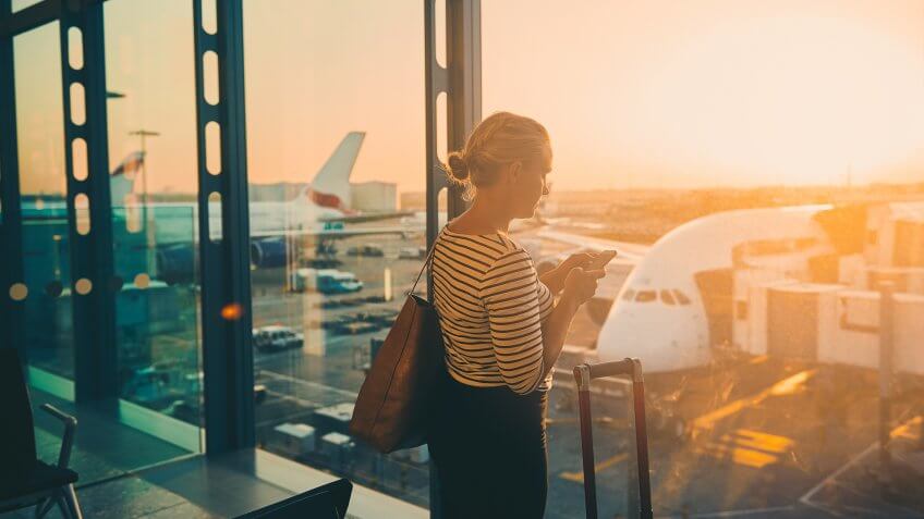 Shot of a young woman using a cellphone while looking through the window of an airport departure lounge.