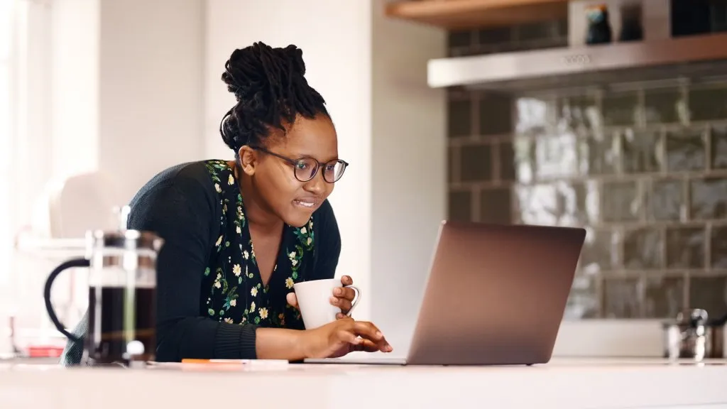 Shot of a young african american woman with a coffee cup using laptop in kitchen at home.