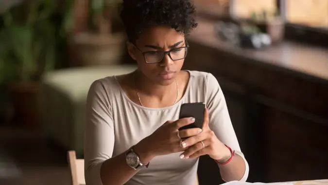 Upset confused african woman holding cellphone having problem with mobile phone, frustrated angry mixed race girl reading bad news in message looking at smartphone annoyed by spam or missed call.