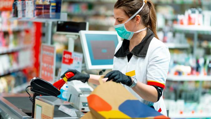 Woman cashier wearing protective face mask and gloves to prevent viruses, scanning disinfection products at the cash register and packing in paper bag.
