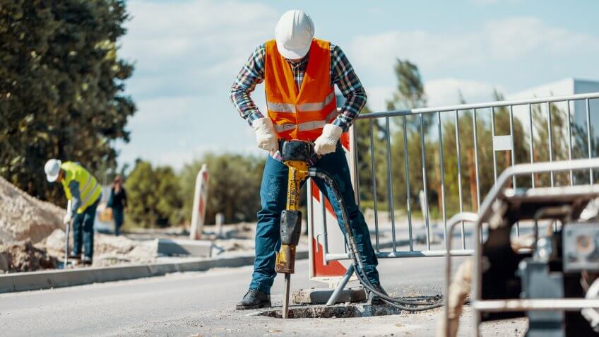 Worker in reflective vest with drill repairing street during roadworks.