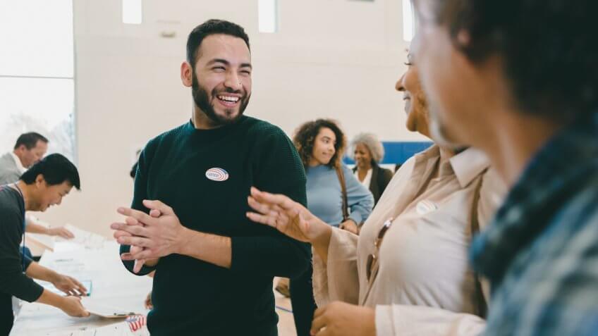 Cheerful mid adult Hispanic man smiles and laughs with friends as they wait to vote on election day.