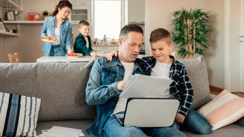 Father Works From Home While His Younger Son Makes Him Company While Mum With Other Son Sets Lunch in the Background.