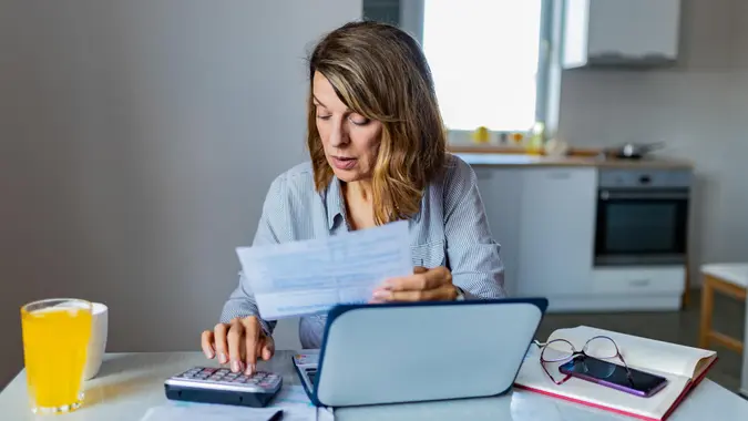 Senior Woman Calculating Domestic Expenses Making Notes, Surrounded With Papers and Gadgets, Sitting at Dining Table.