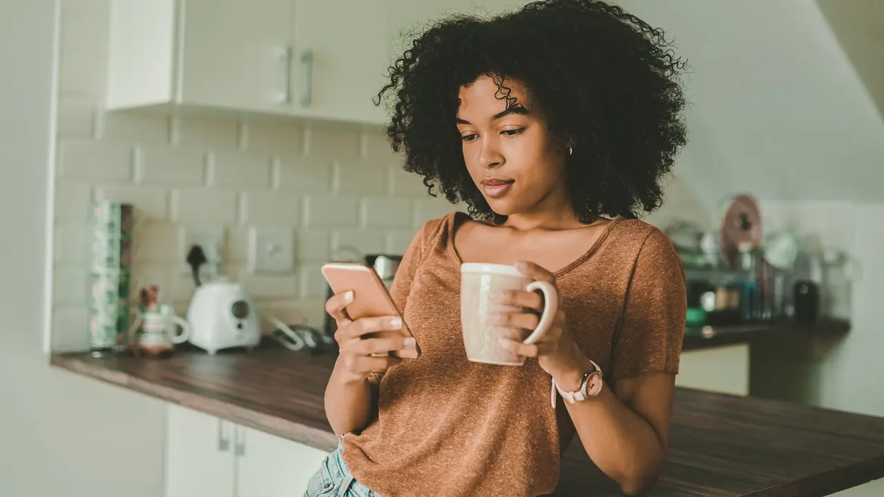 Young woman using a smartphone and having coffee in the kitchen at home.