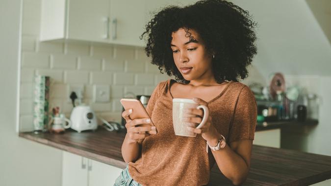 Aufnahme einer jungen Frau, die ein Smartphone benutzt und zu Hause in der Küche Kaffee trinkt.