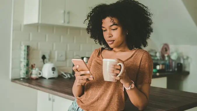 Shot of a young woman using a smartphone and having coffee in the kitchen at home.