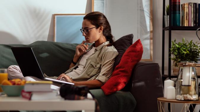 A young woman with glasses using a laptop while lying on a couch in her living room.