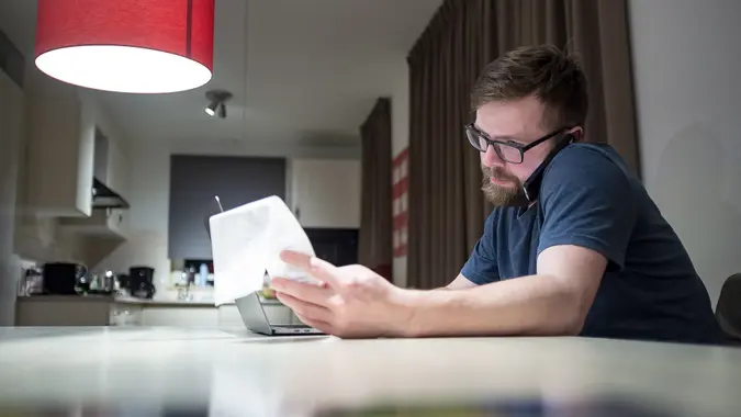 Calm, focused bearded man with glasses manages finances by phone while sitting at home in the kitchen, he looks at the receipt he holds in his hand, next to him is an open laptop.