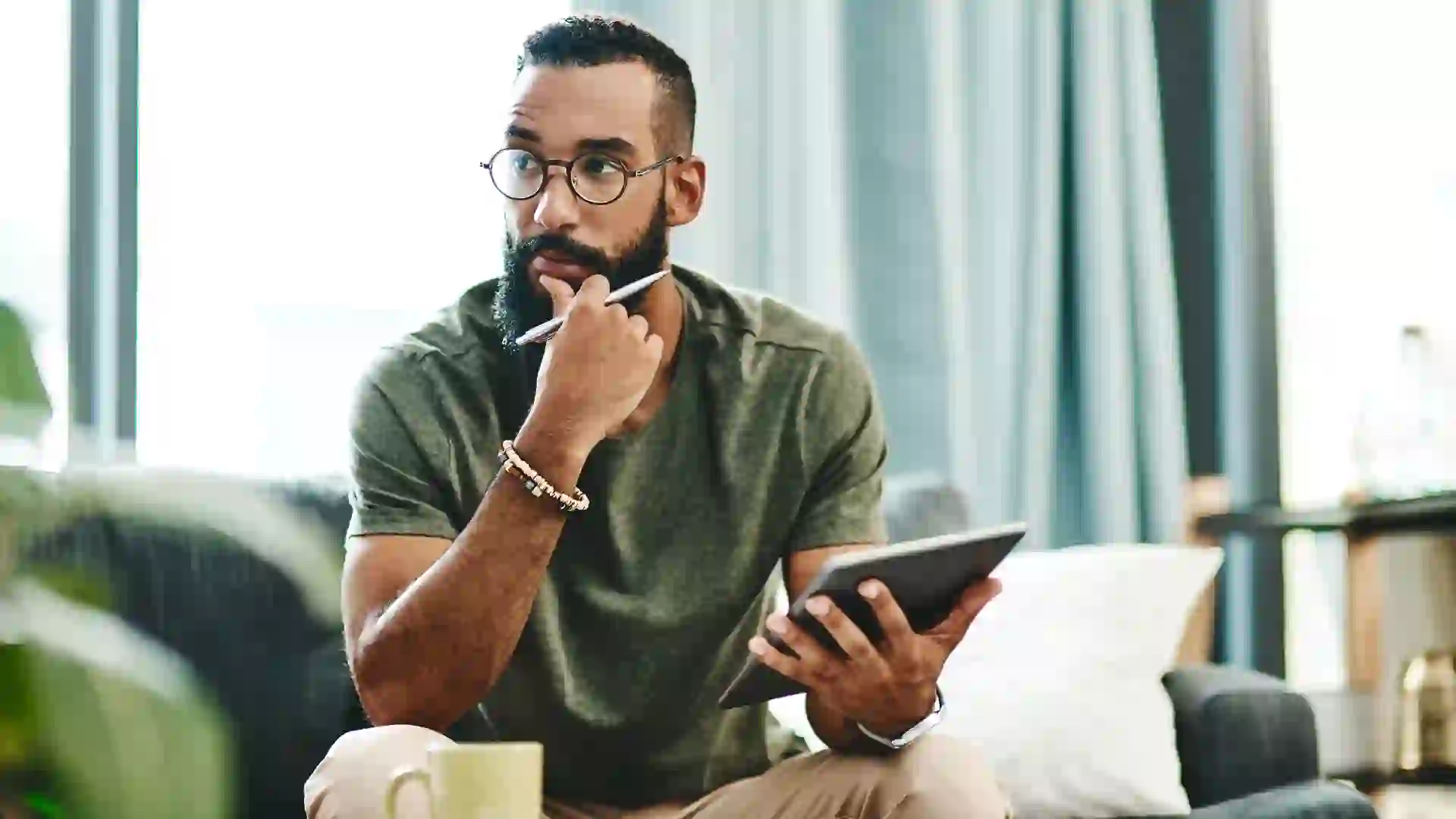 A serious young man concentrates on his finances at home, reviewing numbers on his tablet while sitting in his living room.