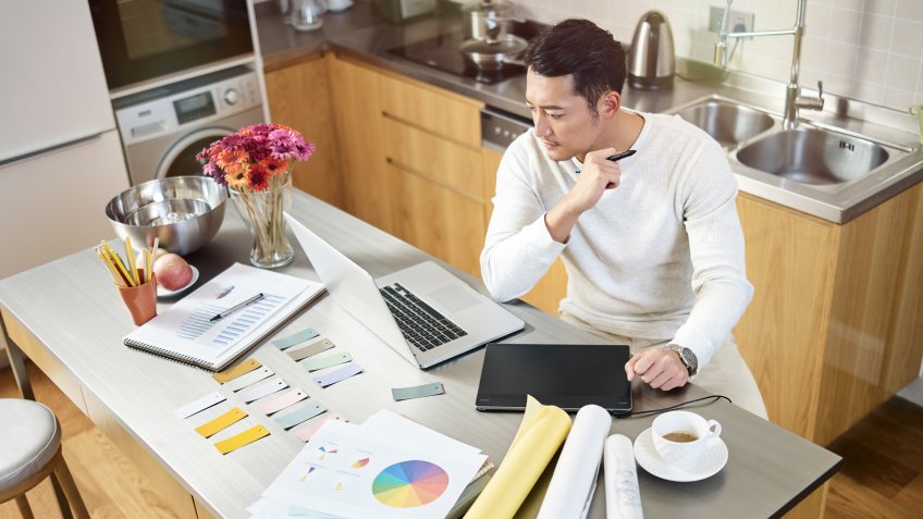 high angle view of a young asian designer sitting at kitchen counter working on a design using laptop computer and digital pen tablet.
