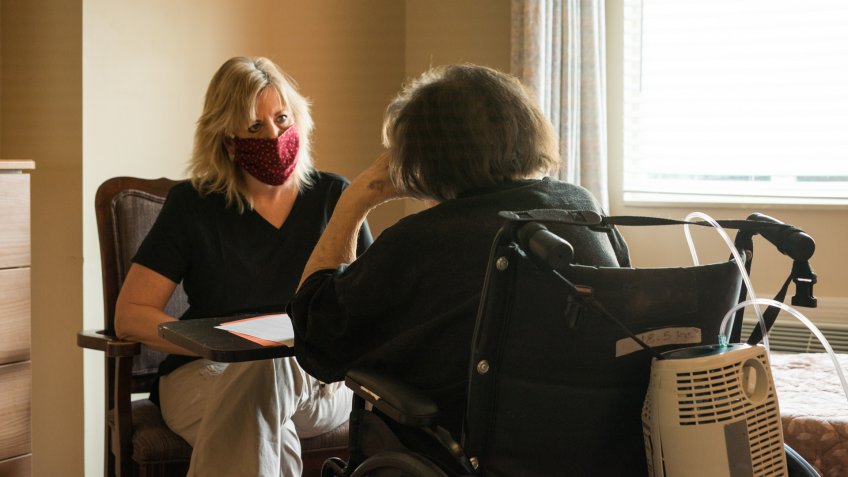 Blond female healthcare worker wearing scrubs, gloves and a red fabric face mask sits across from a patient on oxygen sitting in a wheelchair during a speech therapy session amid the corona virus COVID-19 pandemic.
