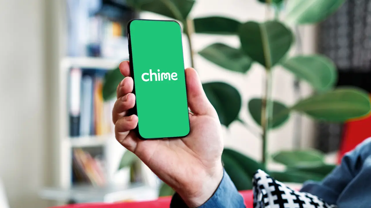 Person holding a phone with the Chime app open in a living room with a bookcase, plant and red furniture in view.