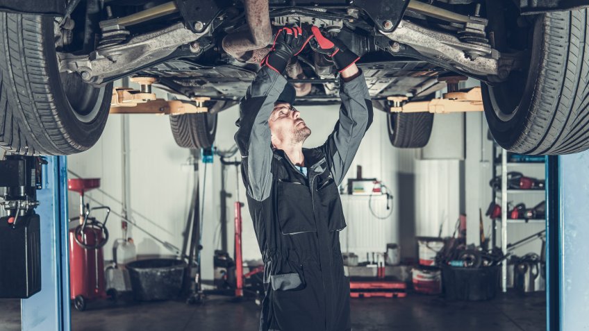 Caucasian Car Mechanic Under Vehicle Looking For Potential Issues with a Drivetrain.
