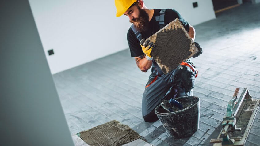 A handsome young bearded man in overalls laying a tile floor.