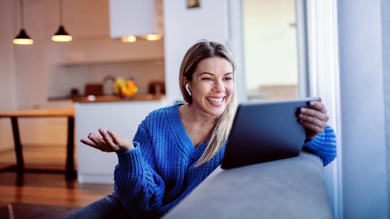 Gorgeous smiling caucasian young blonde in blue sweater and in jeans sitting on sofa and using tablet for video call.