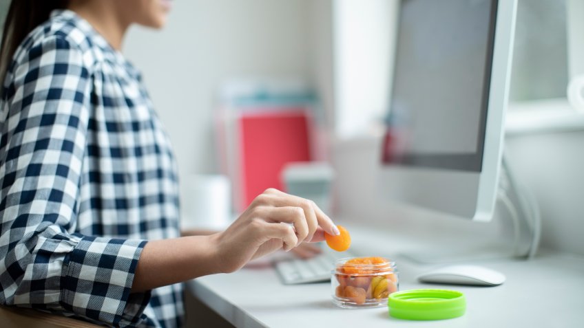 Female Worker In Office Having Healthy Snack Of Dried Apricots At Desk.