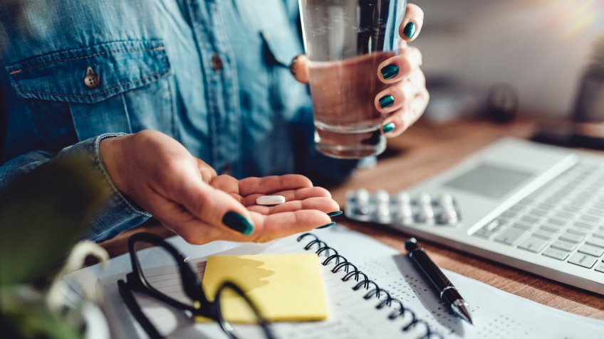 Woman wearing denim shirt sitting by the desk in the office and taking medical pills.