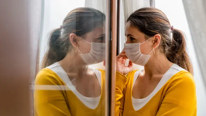 A female coronavirus (Covid-19) or sick patient is forced to self quarantine at her home as she looks through her home window with medical mask to prevent the spread of the virus.