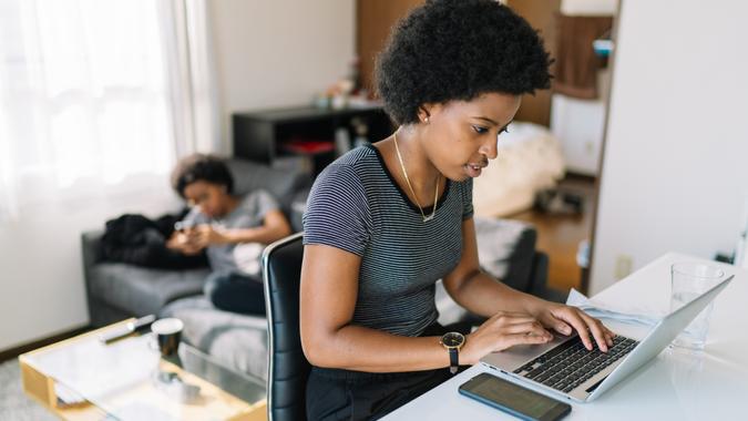 A young and beautiful black woman is using a laptop at home while her identical twin sister is using a a smart phone on the sofa in the background.