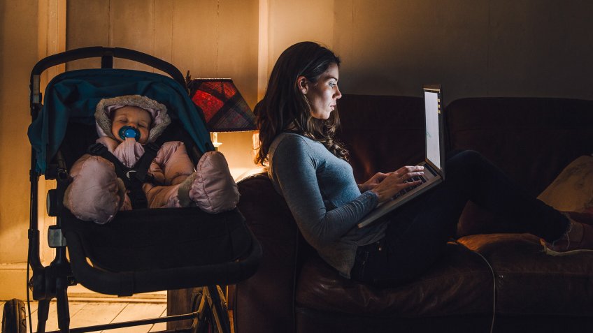 Young mother is working on a laptop on the sofa in her home with her baby daughter sleeping in the pushchair next to her.