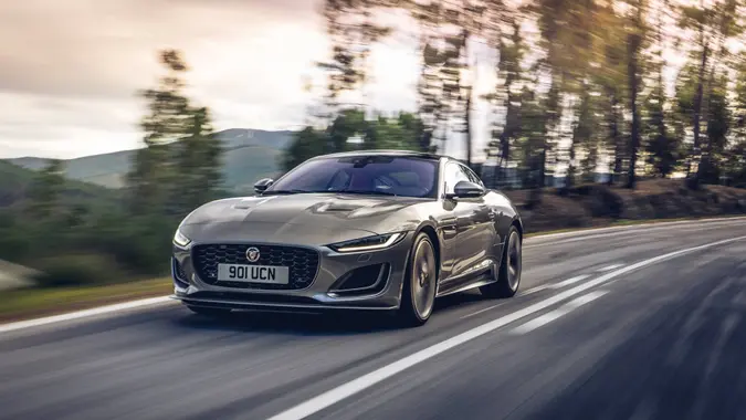 A silver 2021 Jaguar F-Type driving on an open road with trees behind it.
