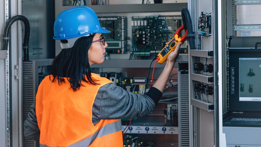 Young woman industrial service electrician engineer wearing protective vest and blue technician helmet testing and checking fridge electric voltage with digital multimeter electric measurement instrument a control panel fuse box in boiler energy control room of modern thermal or nuclear power plant electric energy station.