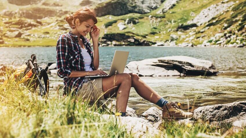 Young woman sitting at a lake side in the mountain and working on a laptop.