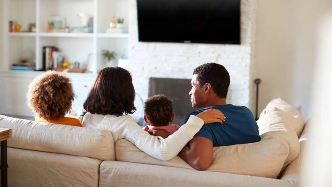 Back view of young family sitting on the sofa and watching TV together in their living room, close up.