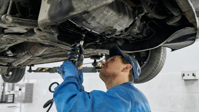 Professional mechanic man in blue uniform working with tools under lifted car in repair service.