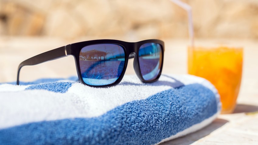 Sunglasses on a towel reflecting blue pool with a drink on the side.