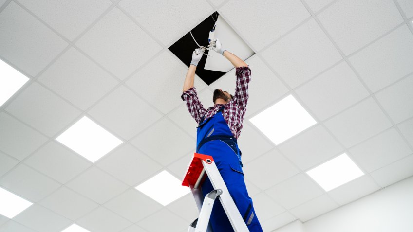 Full Length Portrait Of Electrician On Stepladder Installs Lighting To The Ceiling In Office.