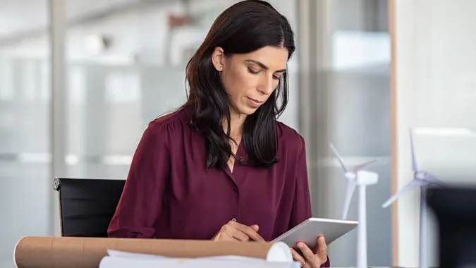 Woman at desk working on architectural plan using digital tablet with scale models of wind turbines.