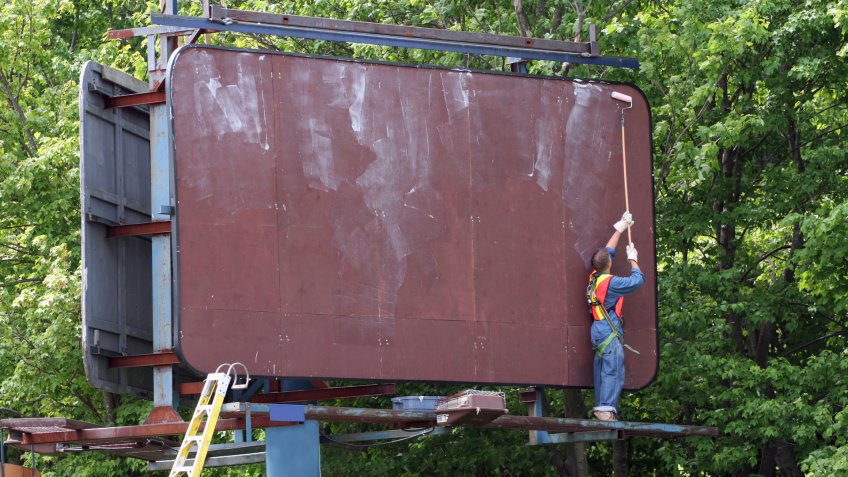 Media sign worker prepares billboard with adhesive prior to installing new advertisement.