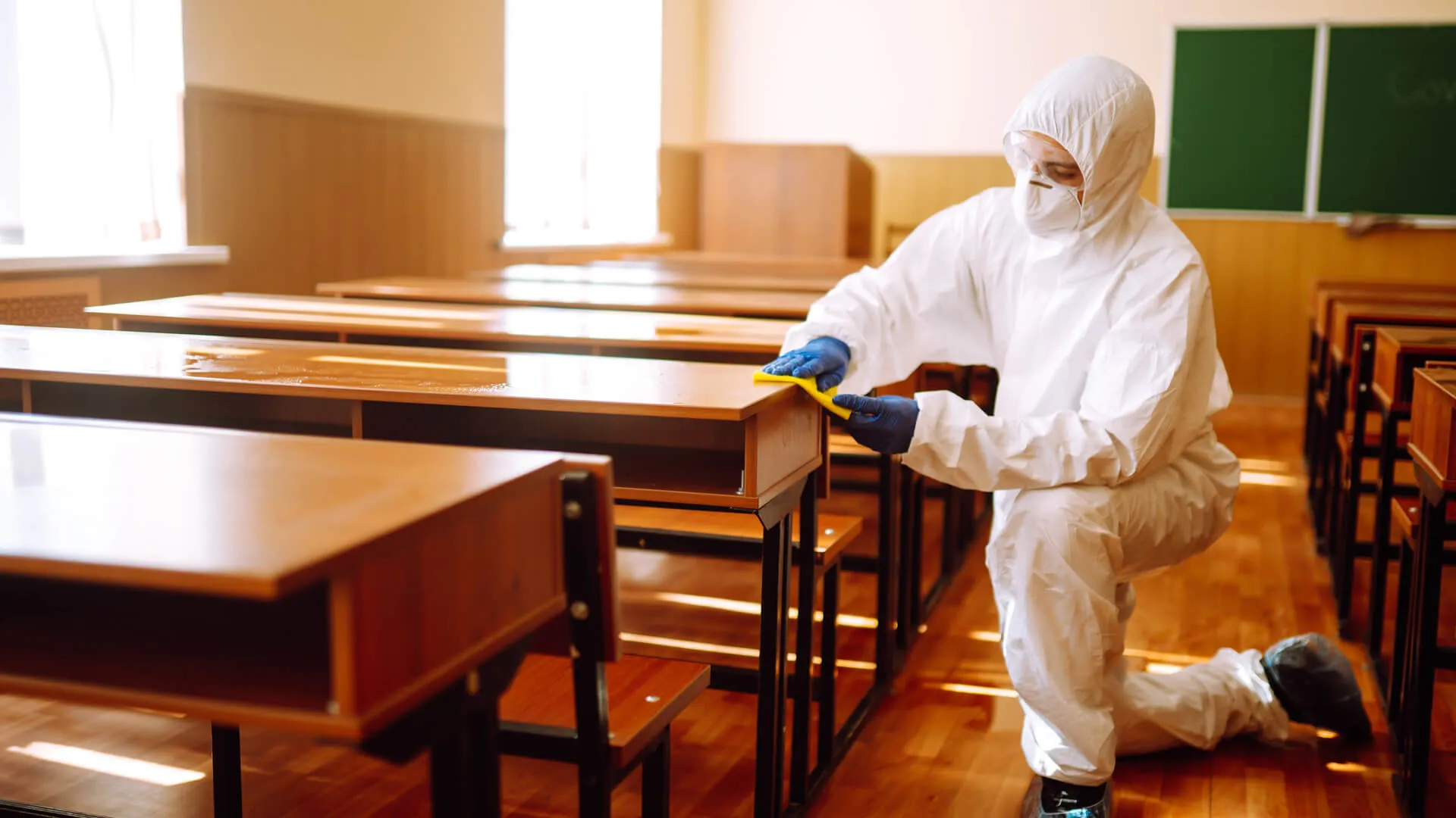 Man in a protective suit washes school desk.
