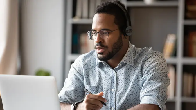 Focused young african businessman wear headphones study online watching webinar podcast on laptop listening learning education course conference calling make notes sit at work desk, elearning concept.