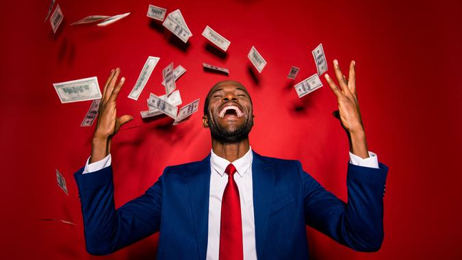 Attractive multiethnic man in style chic classy jacket blue formalwear trendy tux tuxedo stand with closed eyes under money fly rain isolated on red vivid background raised hands up.