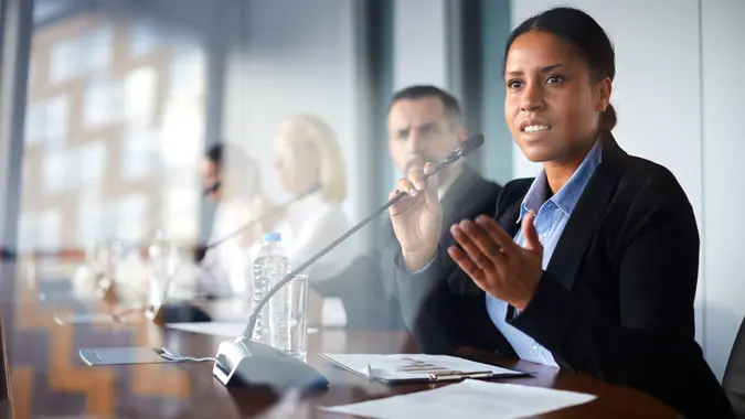 Young female deputee in elegant suit sitting at conference and speaking in microphone to colleague.