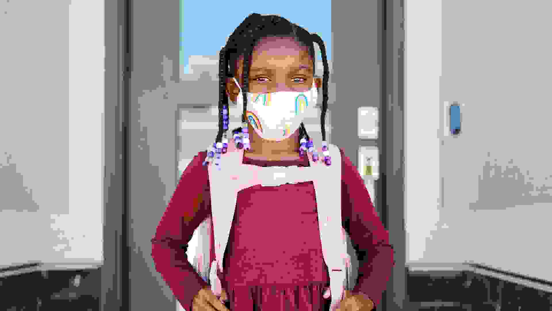 An elementary aged school student leaving her home to go back to school, wearing a mask for protection against infectious disease.