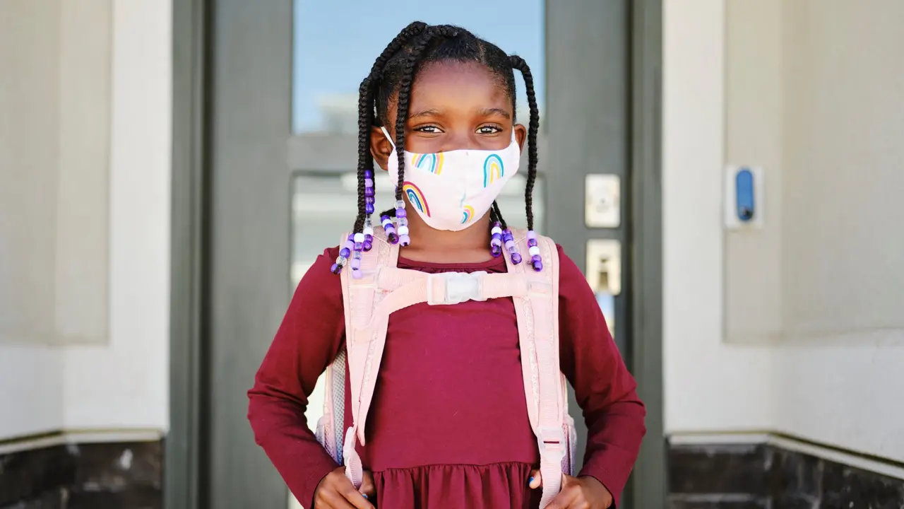 An elementary aged school student leaving her home to go back to school, wearing a mask for protection against infectious disease.