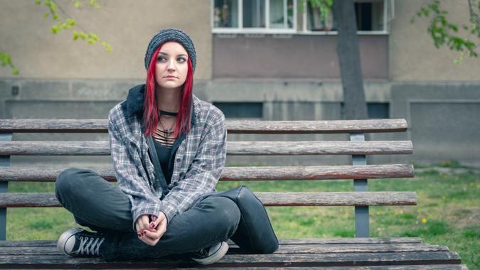 Young beautiful red hair girl sitting alone outdoors on the wooden bench on the street with hat and shirt feeling anxious and depressed after she became a homeless person.