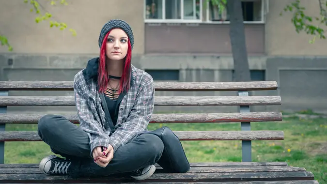 Young beautiful red hair girl sitting alone outdoors on the wooden bench on the street with hat and shirt feeling anxious and depressed after she became a homeless person.