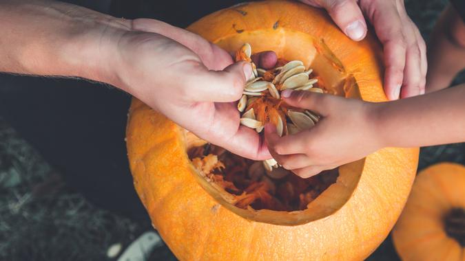 A close up of daughter and father hand who pulls seeds and fibrous material from a pumpkin before carving for Halloween. Prepares jack-o-lantern. Decoration for party. Little helper. Top view.