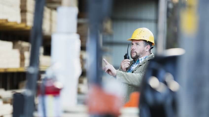 A mid adult Hispanic man in his 30s working in a lumber yard or home improvement store, talking on his walkie-talkie, looking for something on the storage shelf in the warehouse.
