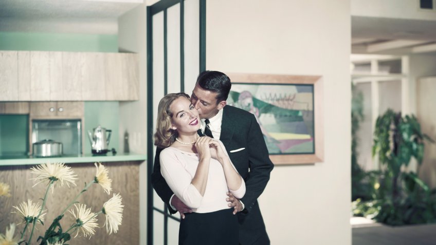1966, Two People, Adults Only, Young Adult, Young Women, Mid Adult, Mid Adult Men, Caucasian Appearance, Indoors, Horizontal, Colour, Photography, Three Quarter Length, Focus On Foreground, Full Suit, Smart, Casual, Blonde Hair, Standing, Kissing, Looking At Camera, Toothy Smile, Cheerful, Hand On Hip, Flower, Living Room, Heterosexual Couple, Togetherness, Happiness, Love, Bonding, Romance, 1966