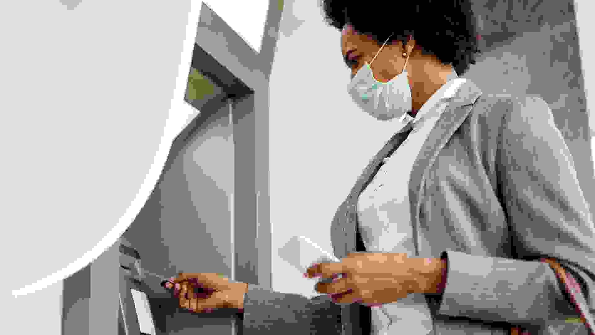 Low angle view of African American businesswoman inserting credit card and withdrawing cash at ATM  while wearing protective mask on her face.